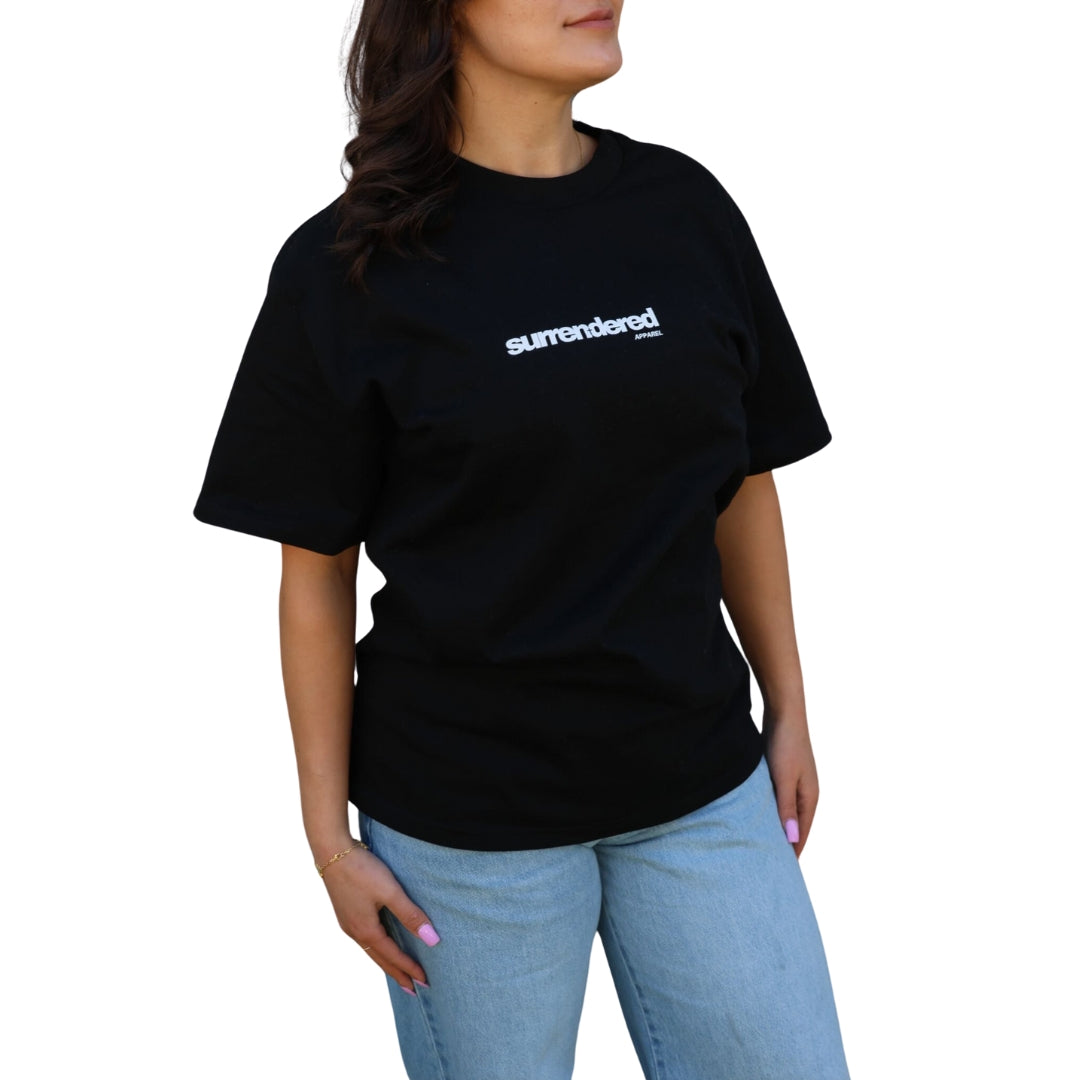 Brunette Woman wearing a Surrendered Apparel Christian unisex Oversize T-shirt in Black. Shirt is thick cotton in black colour, white Surrendered Apparel Logo across the front of the chest. Woman is wearing blue jeans